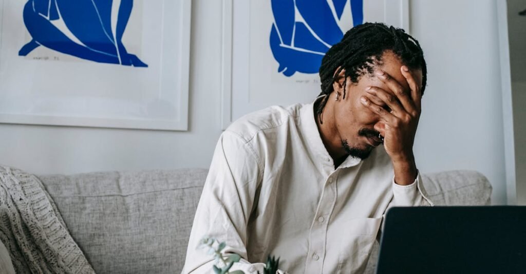 Frustrated young bearded African American male freelancer with dreadlocks in casual shirt working on laptop at home and covering eyes with hand after failure