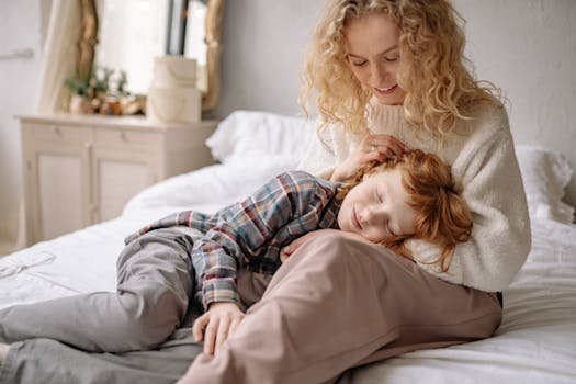 A mother lovingly embraces her son while seated on a bed in a cozy, softly lit room.